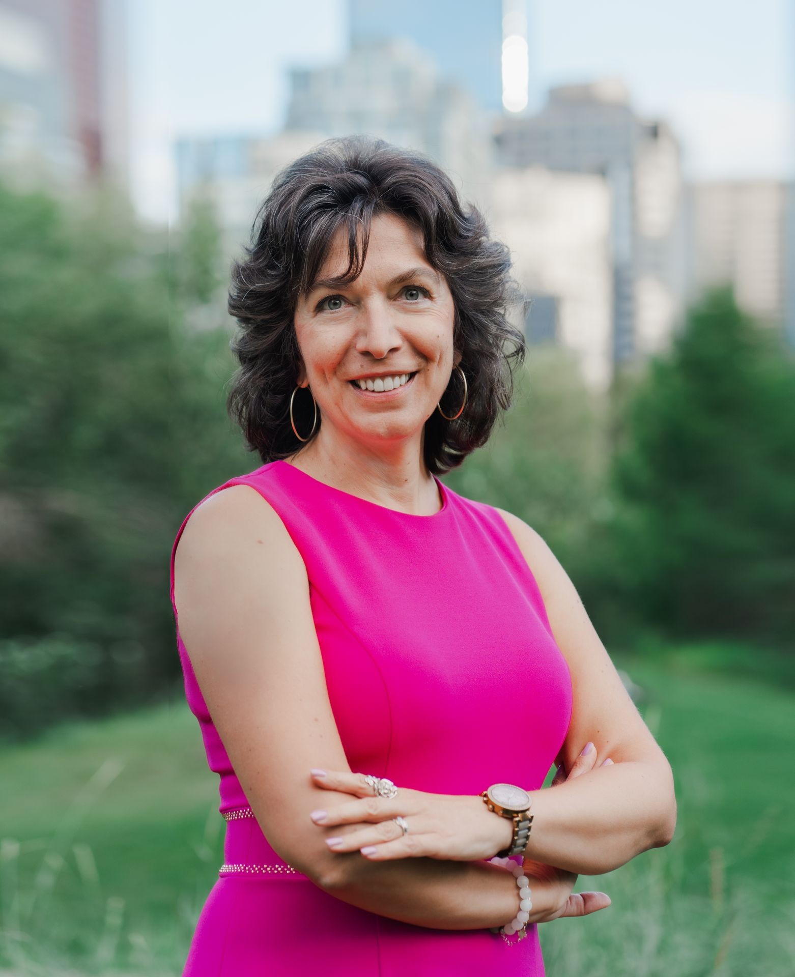 Smiling woman in a bright pink dress standing outdoors with city buildings and greenery in the background.