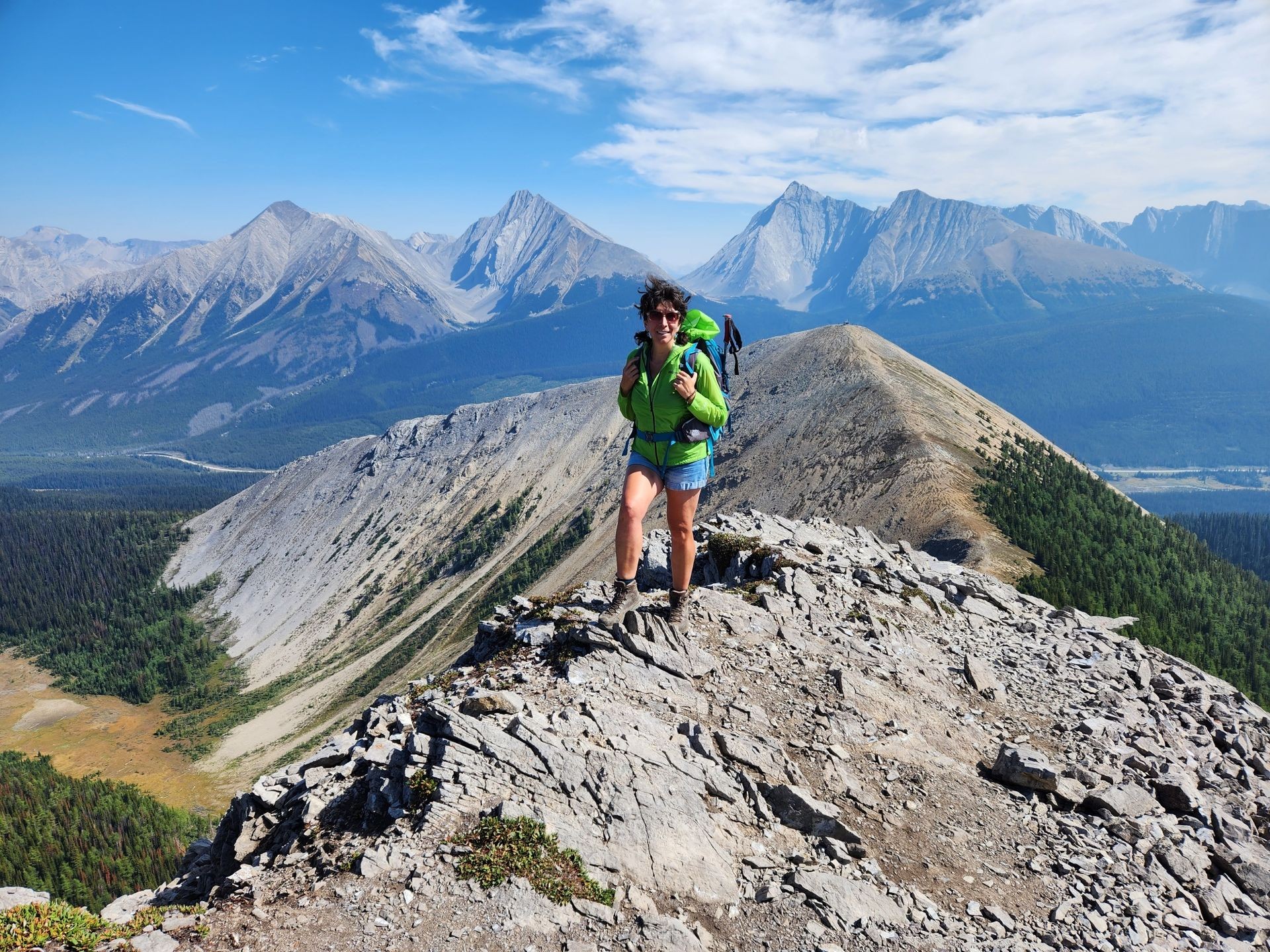 Person hiking on a rocky mountain ridge with a scenic backdrop of rugged peaks and blue sky.